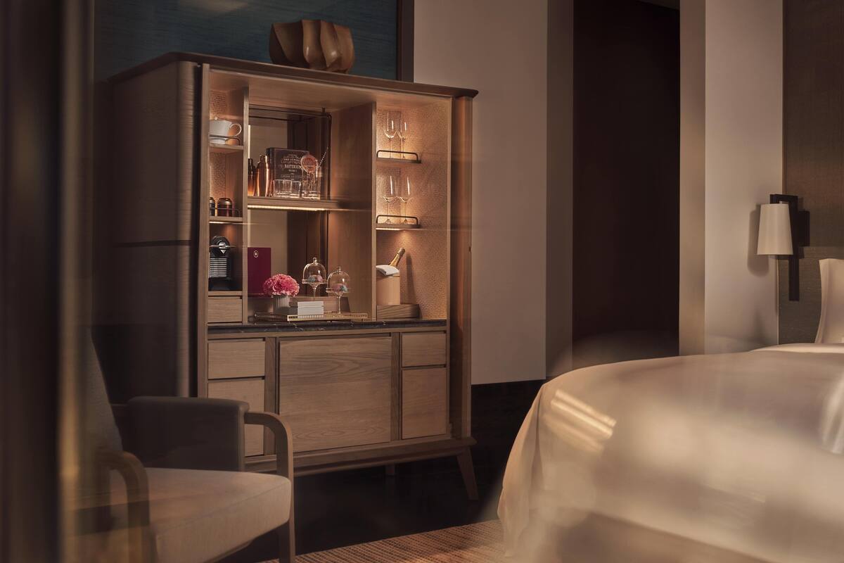 A hotel room with a wooden cabinet containing glassware and a bottle of champagne. There is a bed with a white bedspread and a chair in the corner.