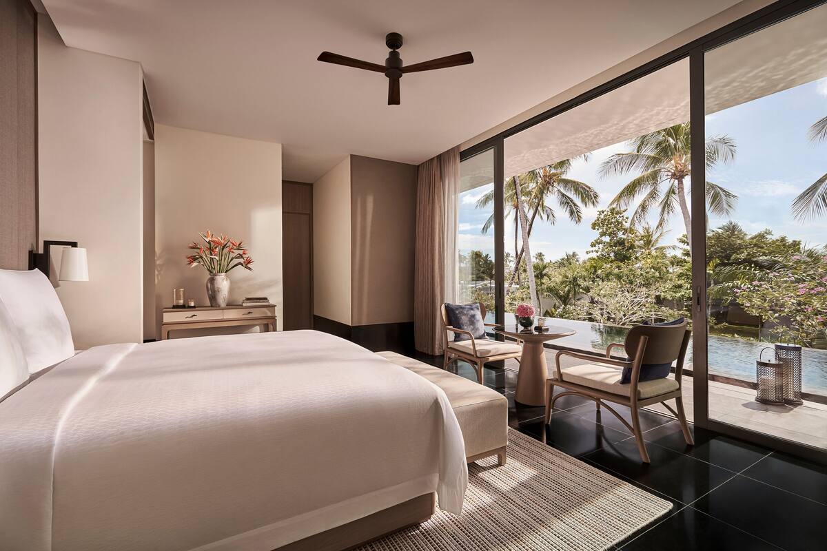 A hotel room with a large bed, a ceiling fan, a nightstand with a vase of flowers, a chair, and a view of a pool and palm trees.