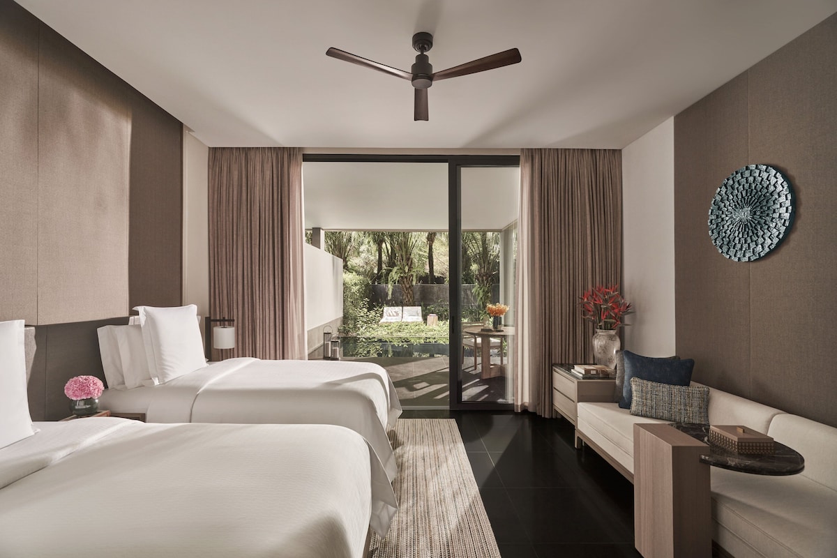 Habitació d'hotel amb dos llits, un ventilador de sostre, vista a un jardí a través d'una porta corredissa i una peça decorativa circular a la paret.