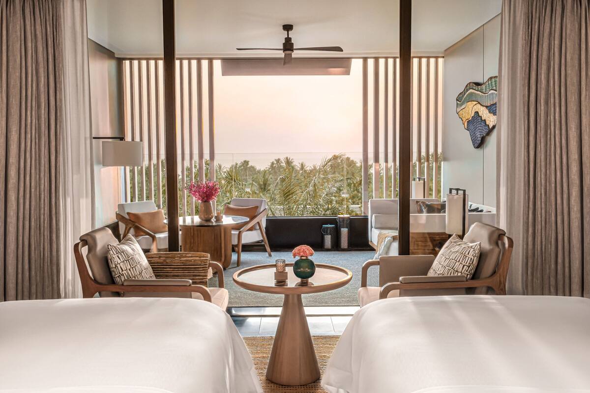 A hotel room with two beds, a round table with a vase, and a balcony with a view of palm trees.
