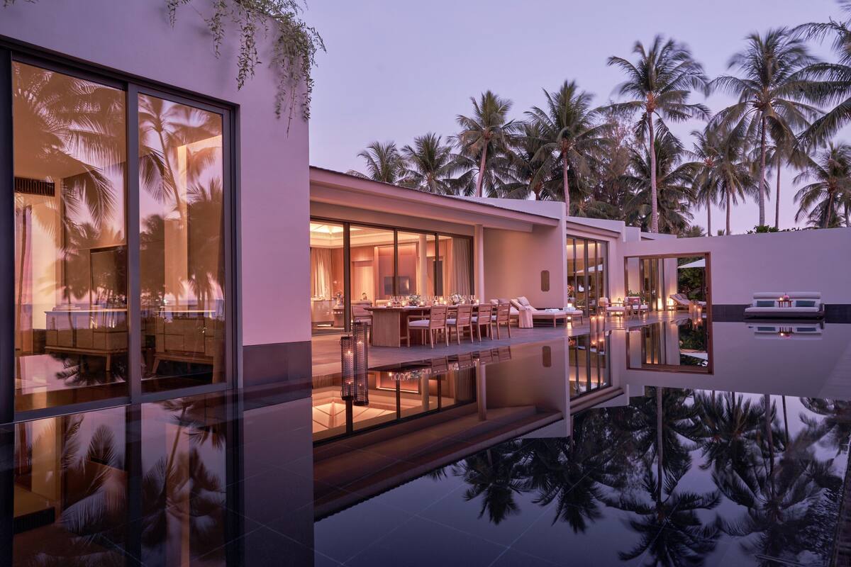 A modern resort with a pool reflecting palm trees, a bar/lounge area, and a dining setup.