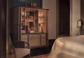 A hotel room with a wooden cabinet containing glassware and a bottle of champagne. There is a bed with a white bedspread and a chair in the corner.