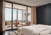 A modern hotel room with a large bed, a balcony with a view of the sea, and a seating area with a table and chairs.