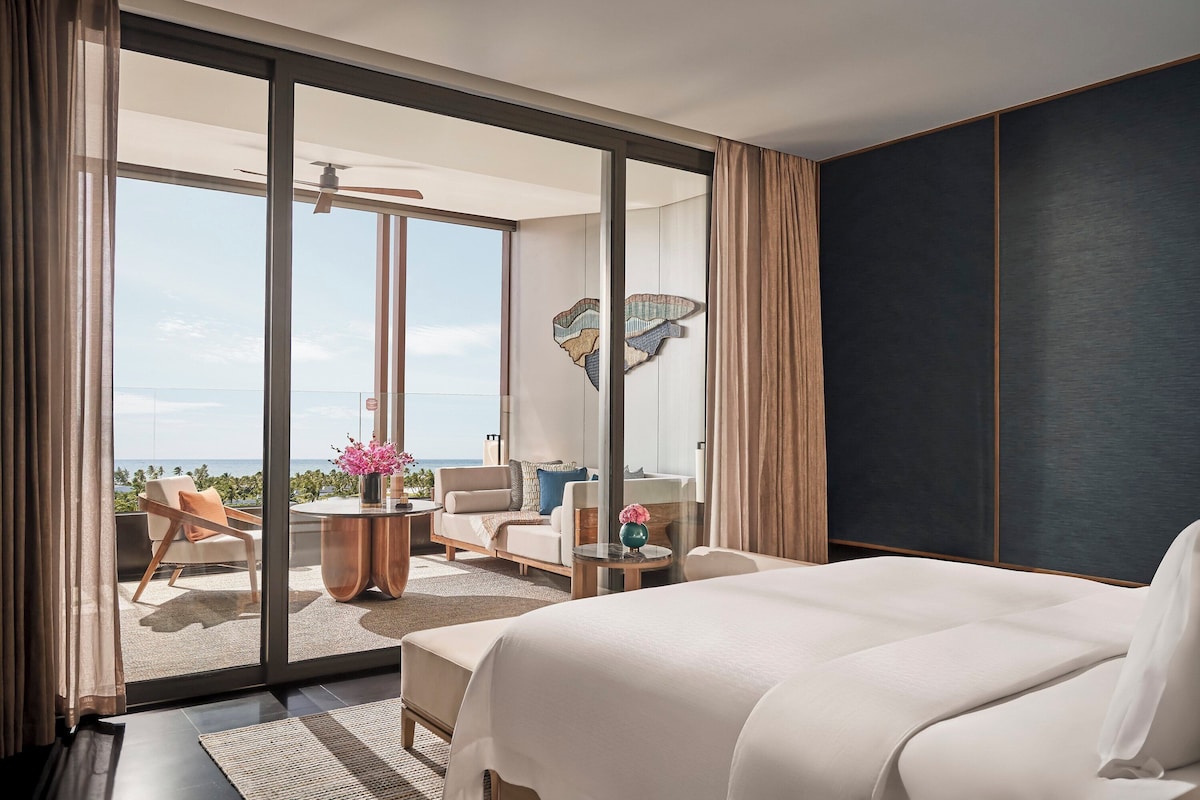 Habitació d'hotel moderna amb un llit gran, un balcó amb vista al mar i una zona de seients amb una taula i cadires.