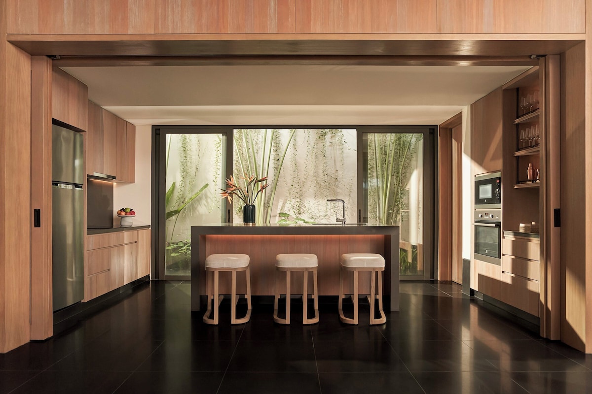 Una cuina moderna amb armaris de fusta, una illa central amb tamborets, una finestra gran amb vista a vegetació i electrodomèstics integrats.