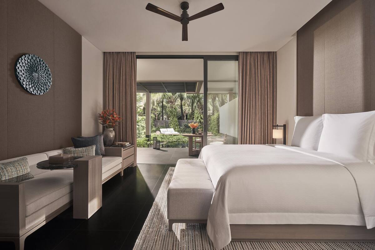 A modern hotel room with a large bed, a sofa, a small table, and a view of the outdoors.