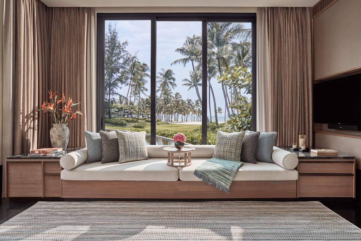 A modern living room with a large window, a sofa with cushions, a small round table with a vase, and a flat-screen TV on a stand.