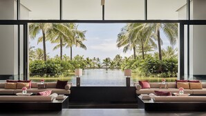 A modern living room with a large window overlooking a pool and palm trees. There are two sofas with cushions and two small tables with lamps. - Regent Phu Quoc (Phu Quoc)