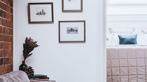 A cozy living room with a sofa, a coffee table, and a bookshelf. There are framed pictures on the wall and a window with a view of a bed. - The Loft (Castlemaine)