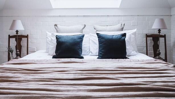 A bedroom with a skylight, a bed with a striped bedspread, two bedside tables with lamps, and a white brick wall. - The Loft (Castlemaine)