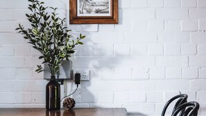 A dining table with chairs, a vase with green foliage, and two framed pictures on a white brick wall. - The Loft (Castlemaine)