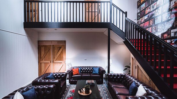 A spacious living area with a dark brown leather sofa set, a coffee table, and a patterned rug. A wooden door and a bookshelf filled with books are visible in the background. - Union Street (Castlemaine)