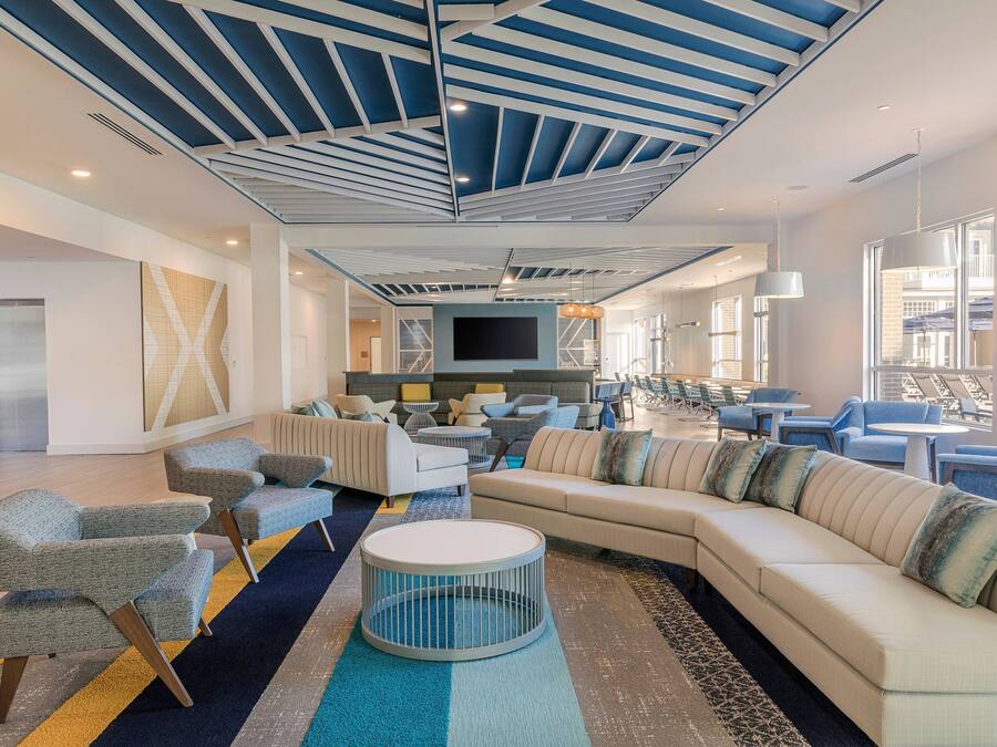 A modern hotel lobby with a large sectional sofa, armchairs, a flat-screen TV, and a ceiling with blue and white geometric patterns.