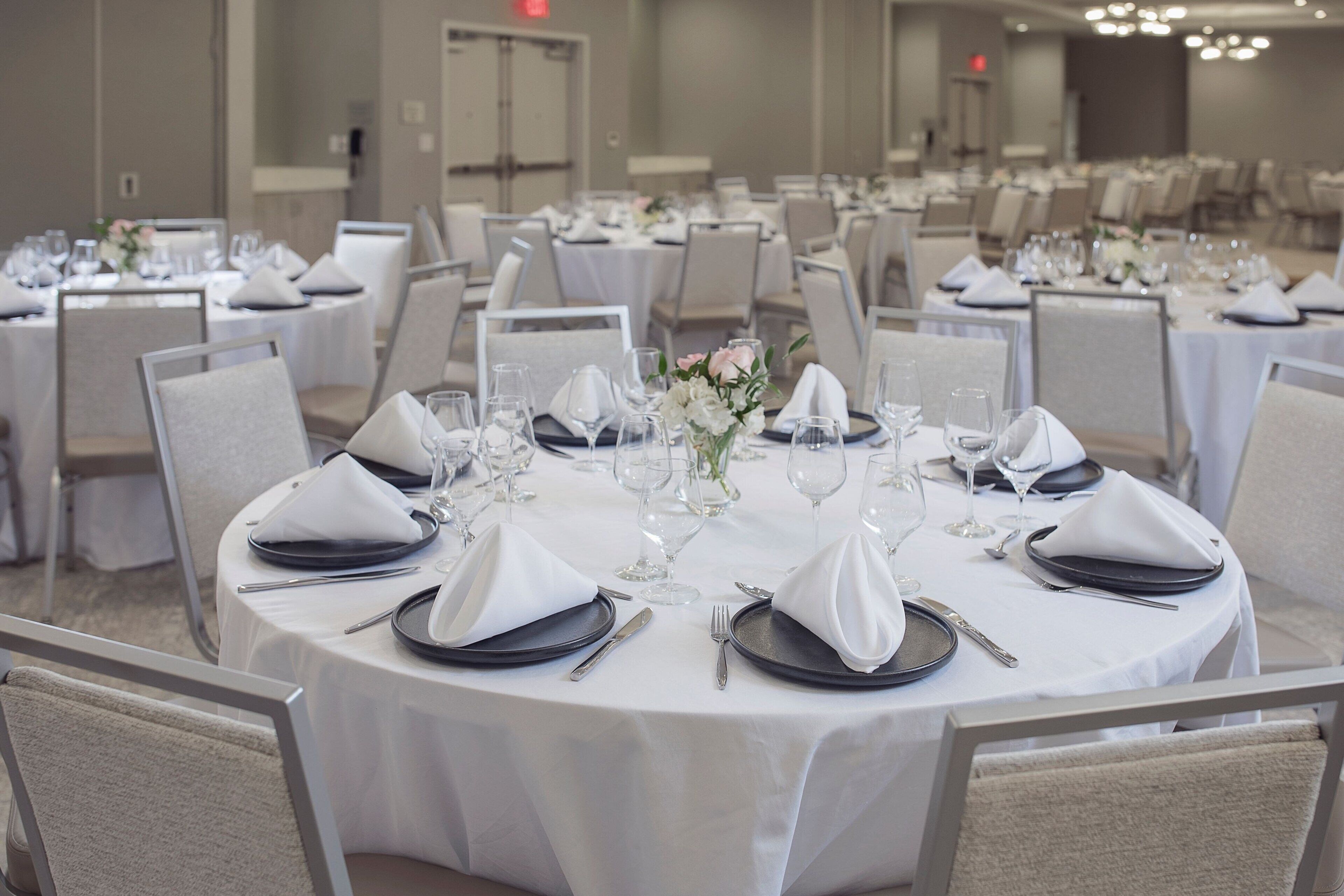 A banquet hall with round tables set for dining, each with white tablecloths, black plates, and silverware.