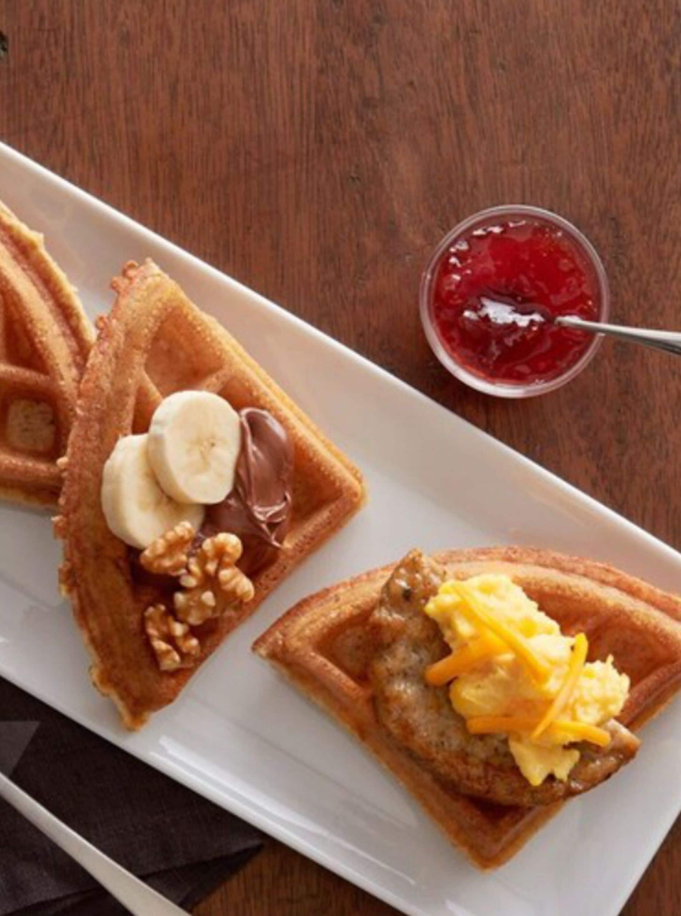 Een wit rechthoekig bord met wafels belegd met bananen, chocolade en walnoten, en een andere wafel met roerei en kaas. Een klein kommetje rode jam is ook zichtbaar.