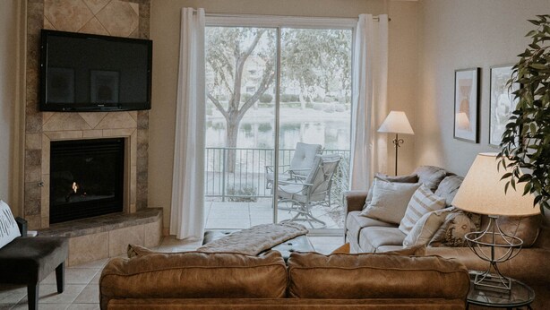 A living room with a fireplace, a sofa, a TV mounted on the wall, and a view of the outdoors through a large window. - Waterfront Mesa Gilbert First Floor Gated Condo Golf Updated Clean Quiet (Mesa)
