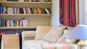 Uma sala de estar aconchegante com um sofá, uma luminária, uma estante repleta de livros e um enfeite pendurado na parede.
