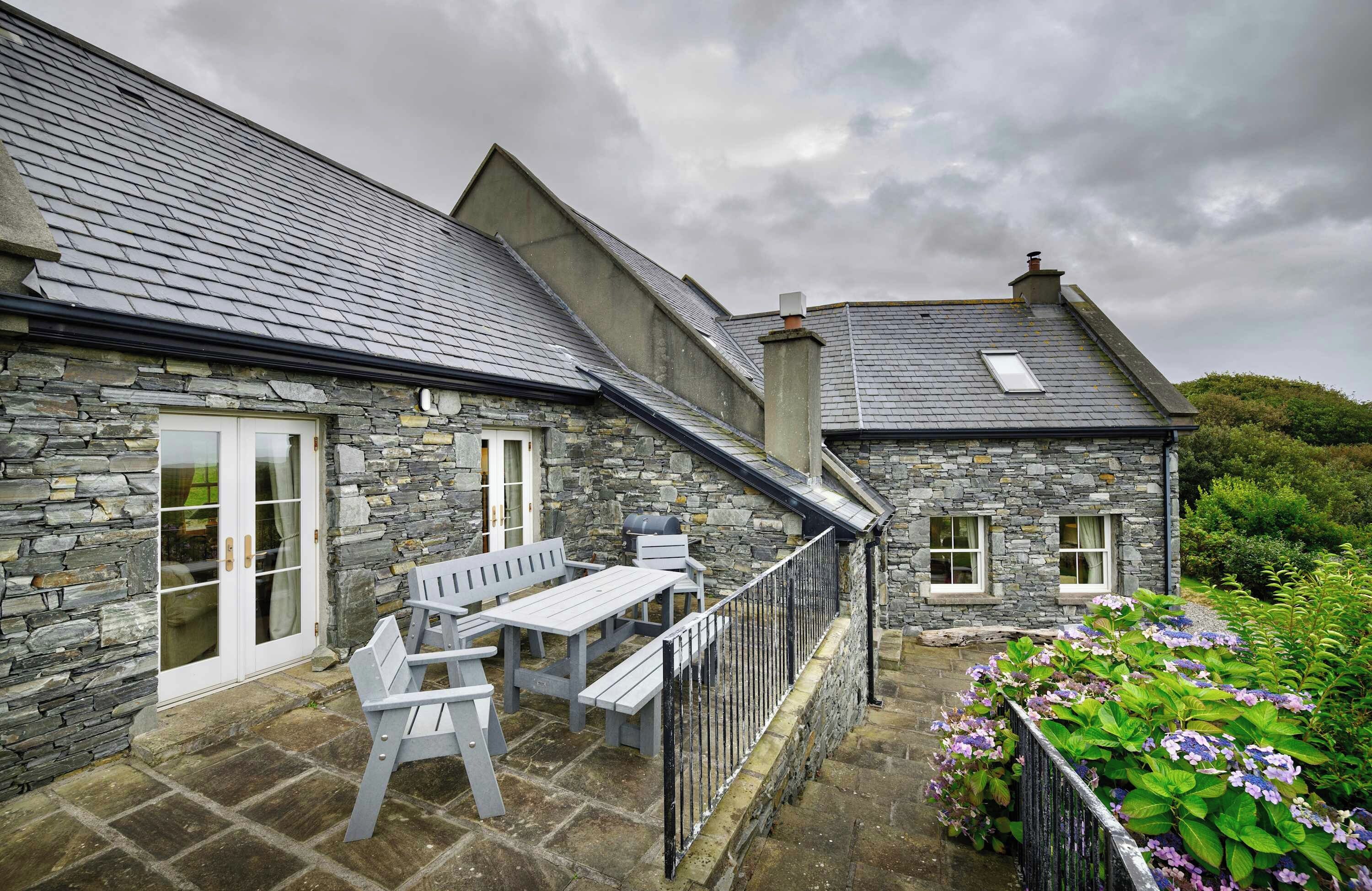 A stone building with a sloped roof, a patio with a table and chairs, and a garden with flowers.