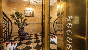 Interior de elevador com piso xadrez, um bonsai em vaso, um candelabro e um quadro na parede.