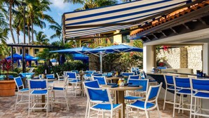 An outdoor patio area with blue and white striped awnings, multiple tables and chairs, and a bar area with a stone wall backdrop. - 2BR Naples Bay Resort Condo (Naples)