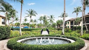 A landscaped area with a circular fountain, palm trees, and a bridge leading to a building. - 2BR Naples Bay Resort Condo (Naples)