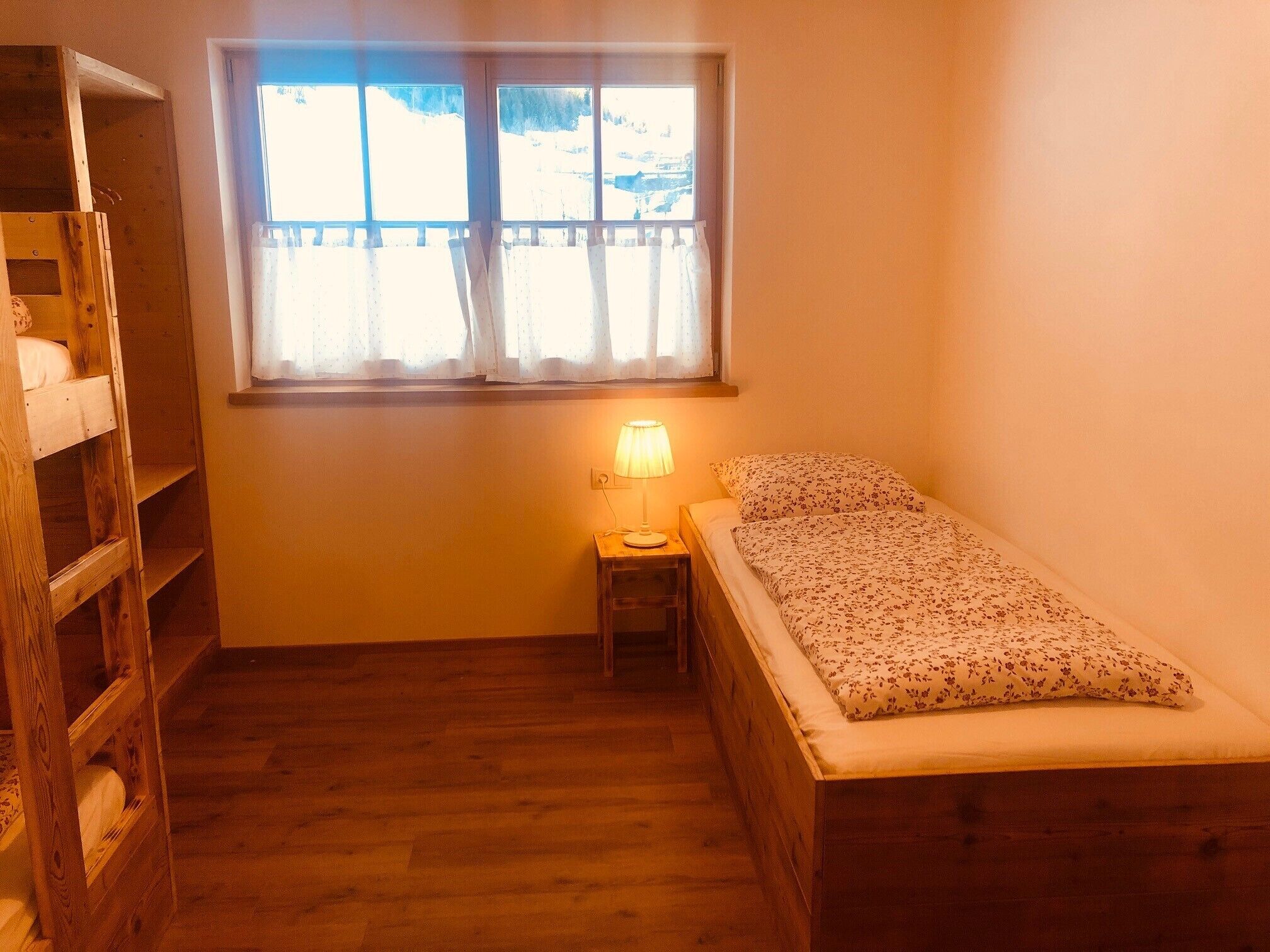 Ein kleiner, gemütlicher Raum mit einem Holzbettkasten, einem Einzelbett mit Blumenmuster-Bettwäsche, einem Nachttisch mit Lampe und einem Fenster mit Blick auf eine verschneite Landschaft.