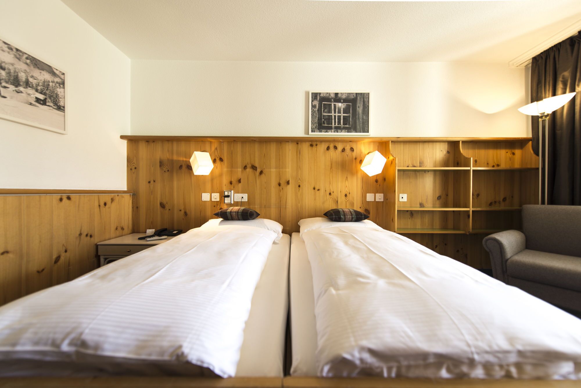 A hotel room with two single beds, wooden paneling on the walls, a small window, and a chair.
