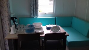 A small kitchenette with a teal-colored bench, a table with a blue container, white plates, and a white bowl. - Mobile home with large terrace 300m from the city (Aire-sur-l'Adour)