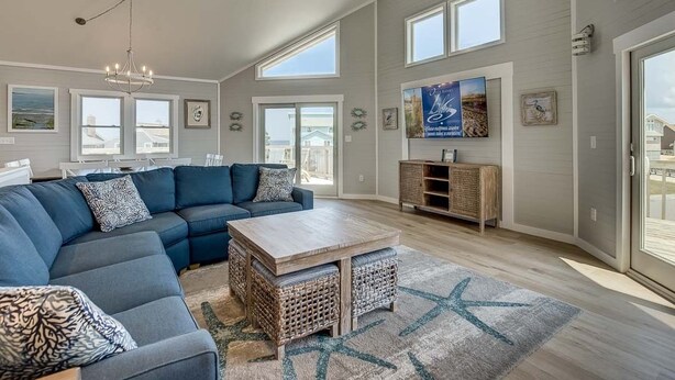 A spacious living room with a large sectional sofa, a wooden coffee table, and a starfish rug. There are large windows, a ceiling fan, and a TV mounted on the wall. - Singing Sandpipers | 325 ft to the beach | Private Pool, Dog Friendly, Community Pool (Duck)