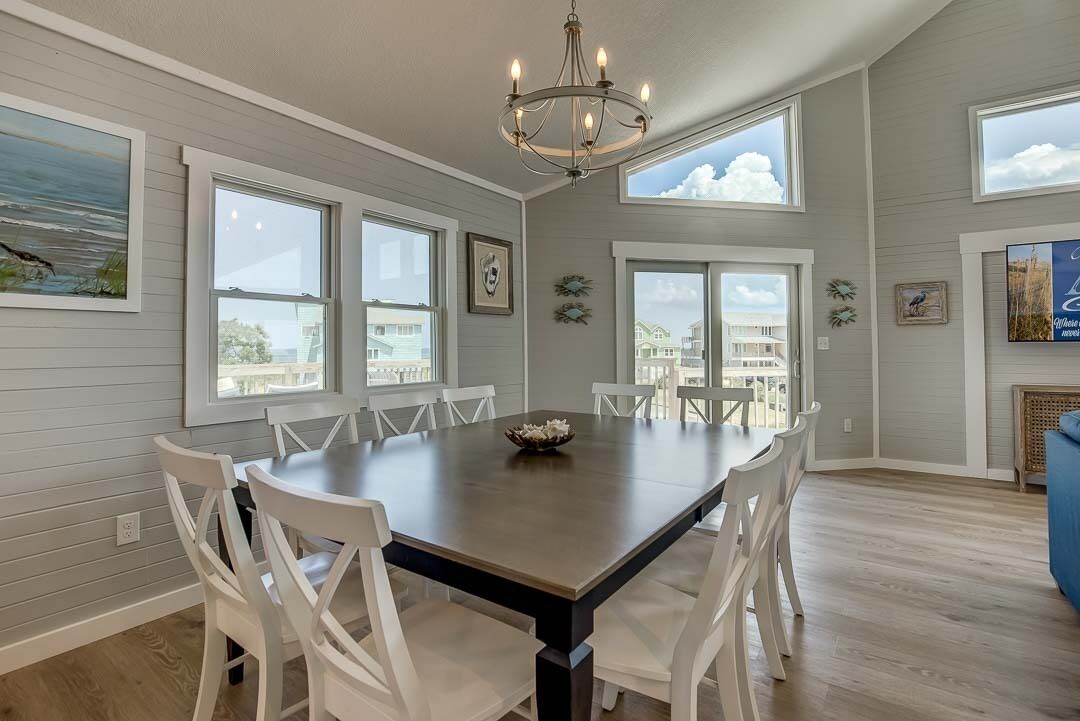 A dining area with a large table and white chairs, large windows with views, and a chandelier.