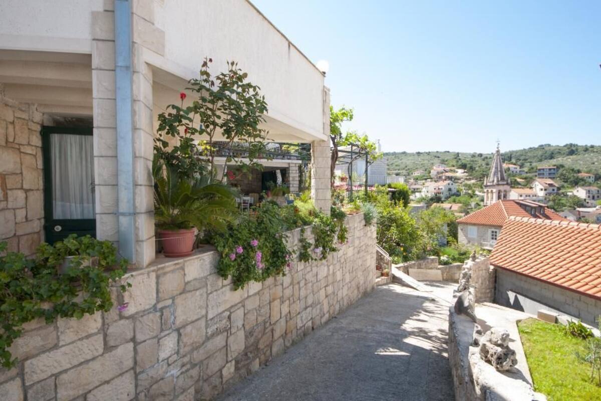 A narrow stone pathway leading to a building with a tiled roof, surrounded by greenery and a view of a town with a church spire.