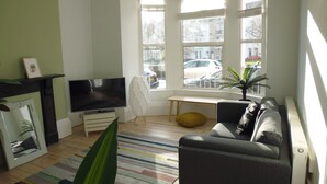 Een woonkamer met een televisie, een bank, een hocker en een groot raam met uitzicht op de straat.