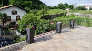 Villa met balkon, omgeven door een metalen hek, met uitzicht op een groen landschap met bomen en een heuvel op de achtergrond.