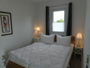 Ein ordentlich bezogenes Bett mit weißer Bettwäsche, zwei Nachttische mit Lampen, ein Fenster mit Vorhängen und ein gerahmtes Bild an der Wand.