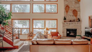 A spacious living room with a stone fireplace, large windows, a staircase, and a leather sofa.