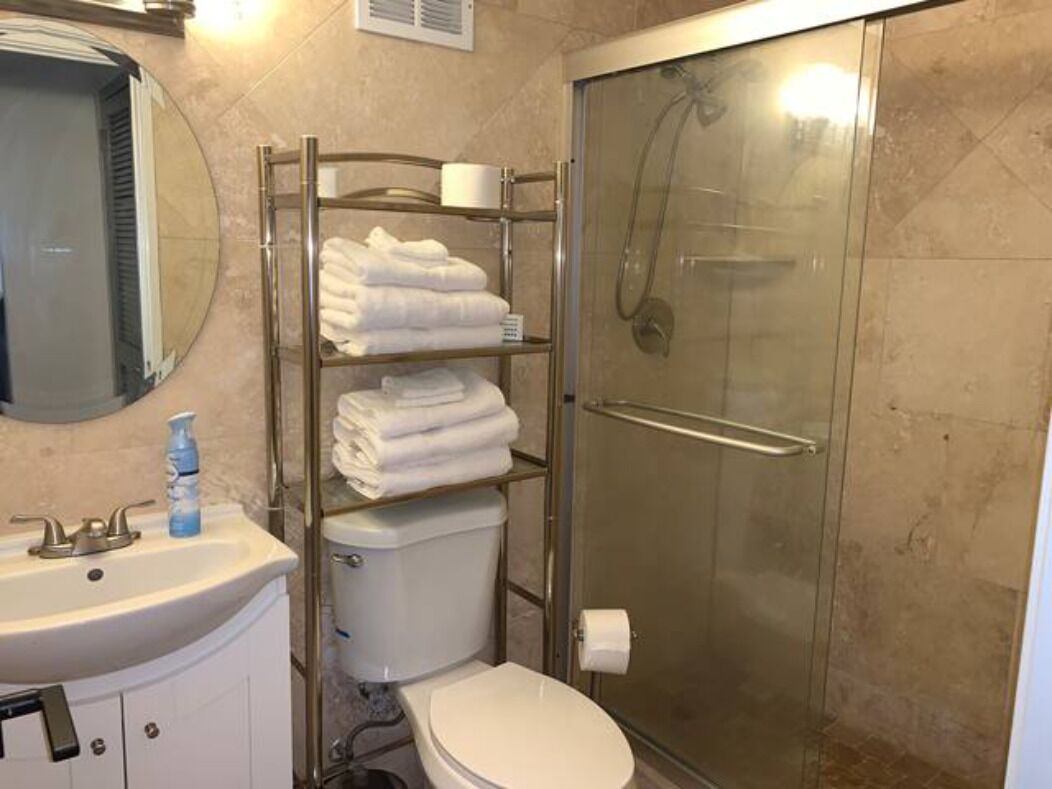 Et badeværelse med et glasafskærmet brusebad, toilet, vask med spejl og et håndklædestativ med sammenfoldede håndklæder.