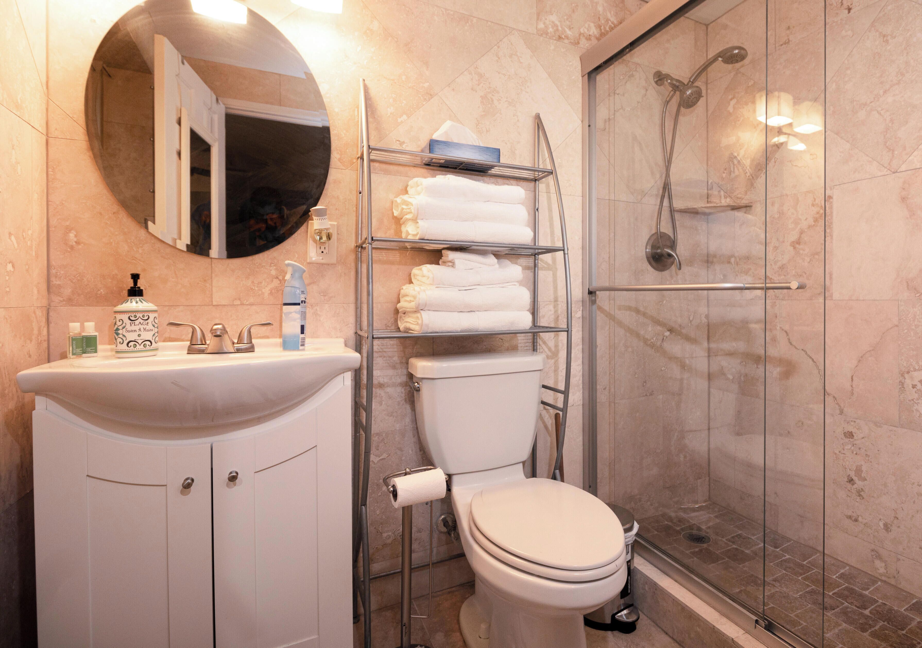 A modern bathroom with a round mirror, a sink with toiletries, a toilet, and a glass-enclosed shower.