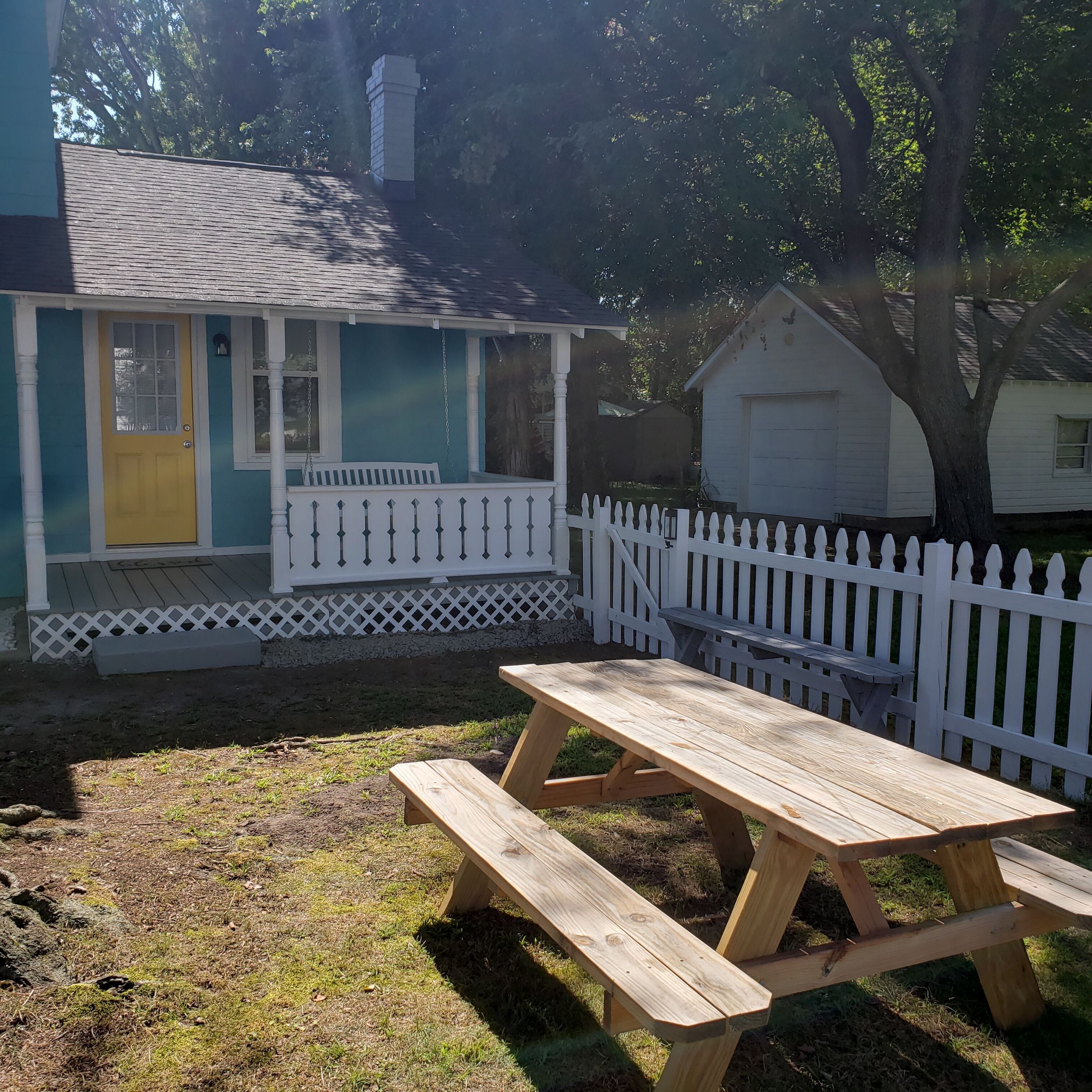 Une maison avec une véranda, une table de pique-nique en bois et une clôture blanche faite de piquets.