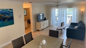 Een ruime woonkamer met een eettafel, een bank en een televisie. Er hangt een schilderij aan de muur en er is een plafondventilator.