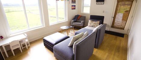 A modern living room with a sofa, armchair, and a small table. There are large windows with a view of a green field.