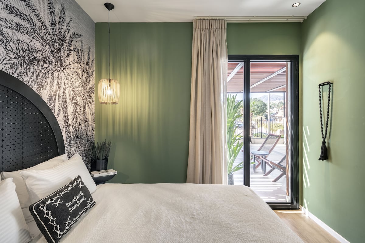 Une chambre à coucher comprenant un lit, une table de chevet, une lampe, un rideau et donnant sur une terrasse extérieure.