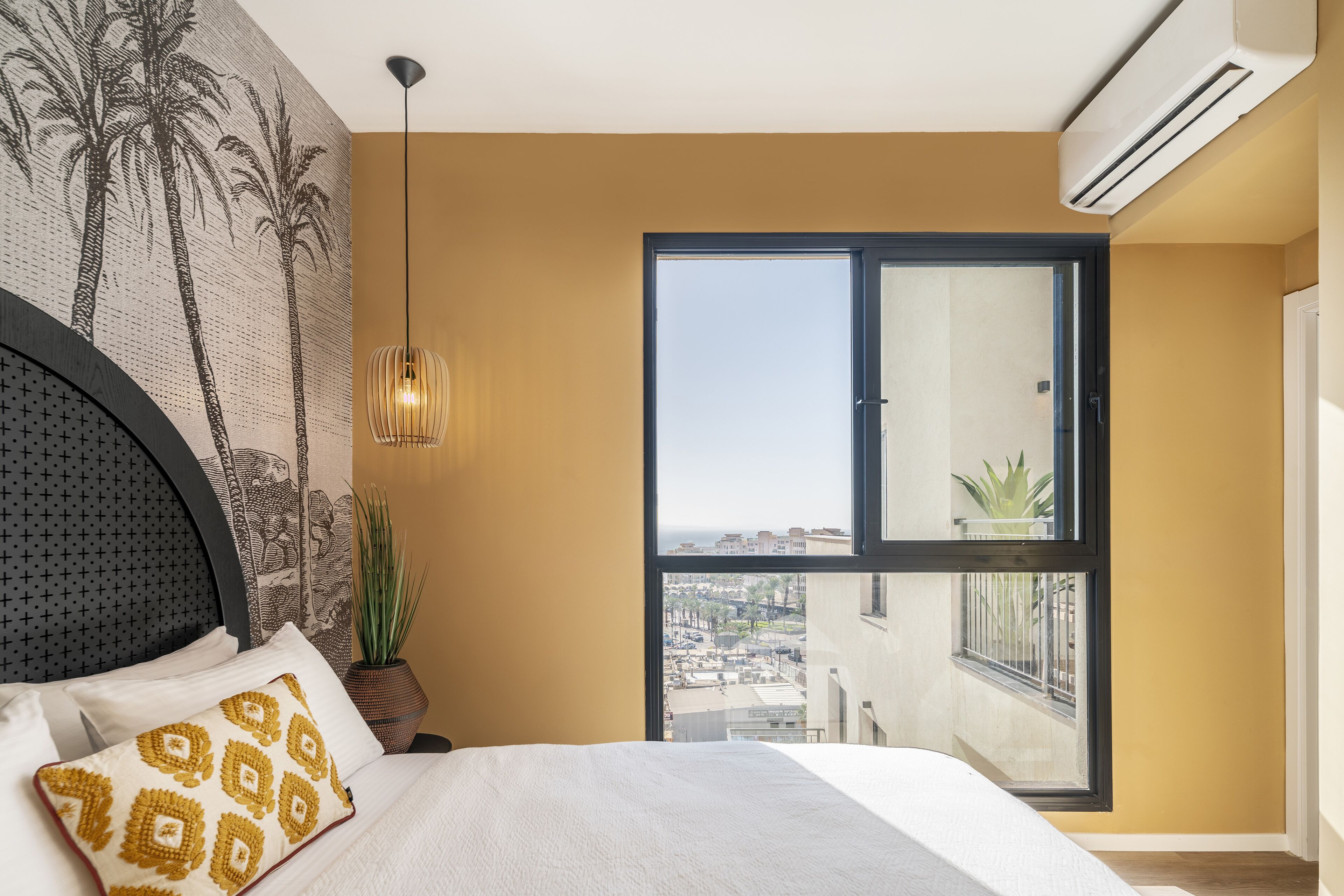 Une chambre avec un grand lit, une vue sur la ville par la fenêtre, une plante et un appareil de climatisation.