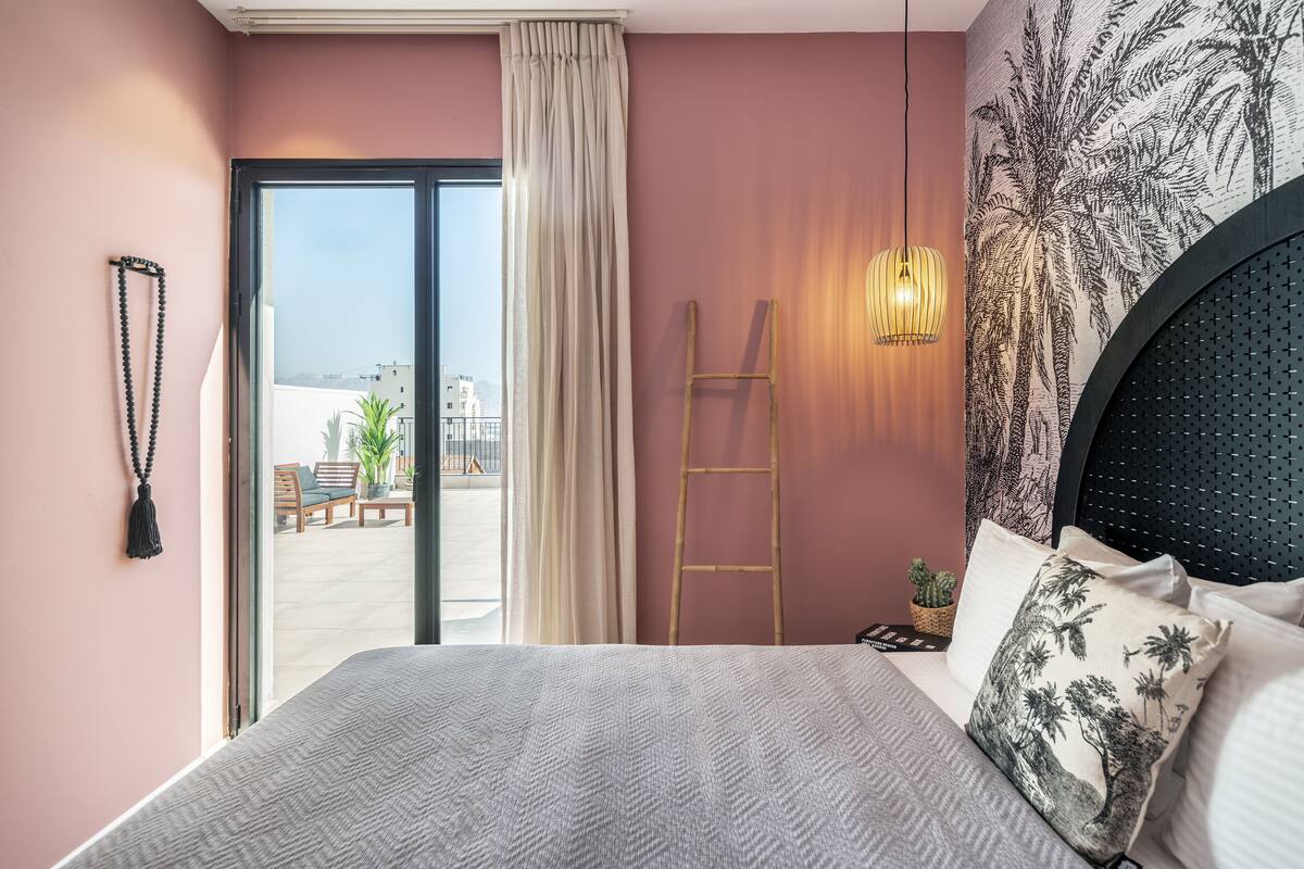 Une chambre avec un grand lit, une vue sur l’extérieur grâce à une porte coulissante, une échelle et un mur orné d’une fresque représentant un palmier.