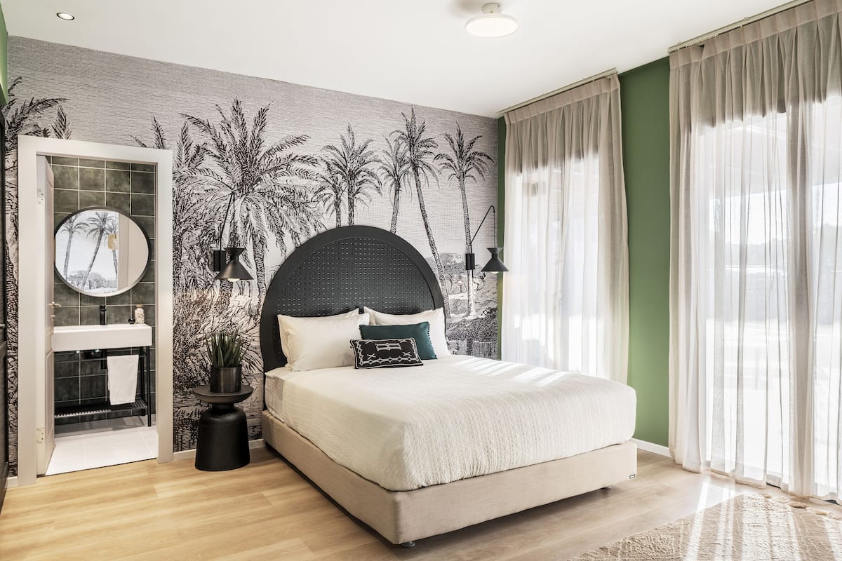 Une chambre avec un grand lit, un miroir rond, une étagère intégrée et une fresque murale représentant des palmiers.