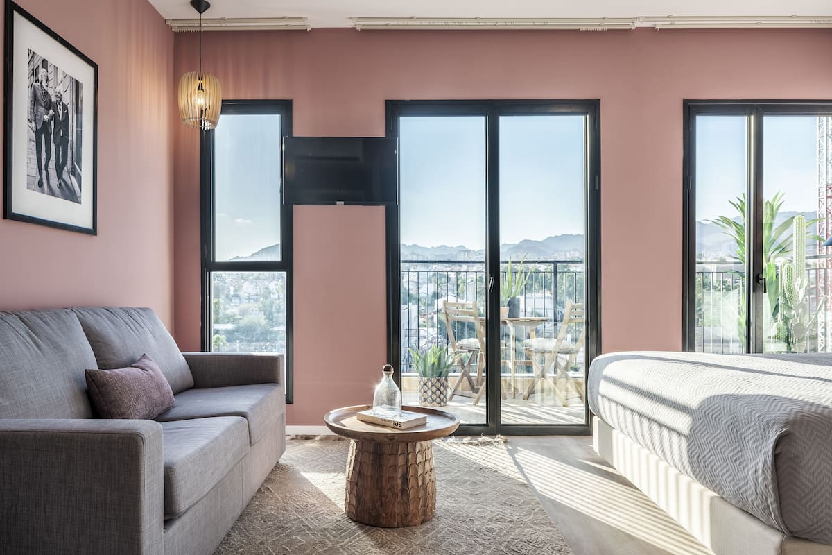 Une chambre moderne avec un canapé, un lit, une table ronde en bois et une grande fenêtre offrant une vue sur la ville et les montagnes.