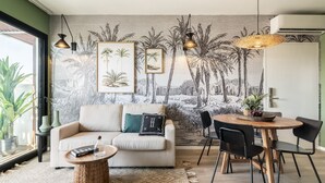 Un salon comprenant un canapé, une table basse et un coin repas avec des chaises. Un mur est décoré d’une fresque représentant des palmiers et une scène de plage.