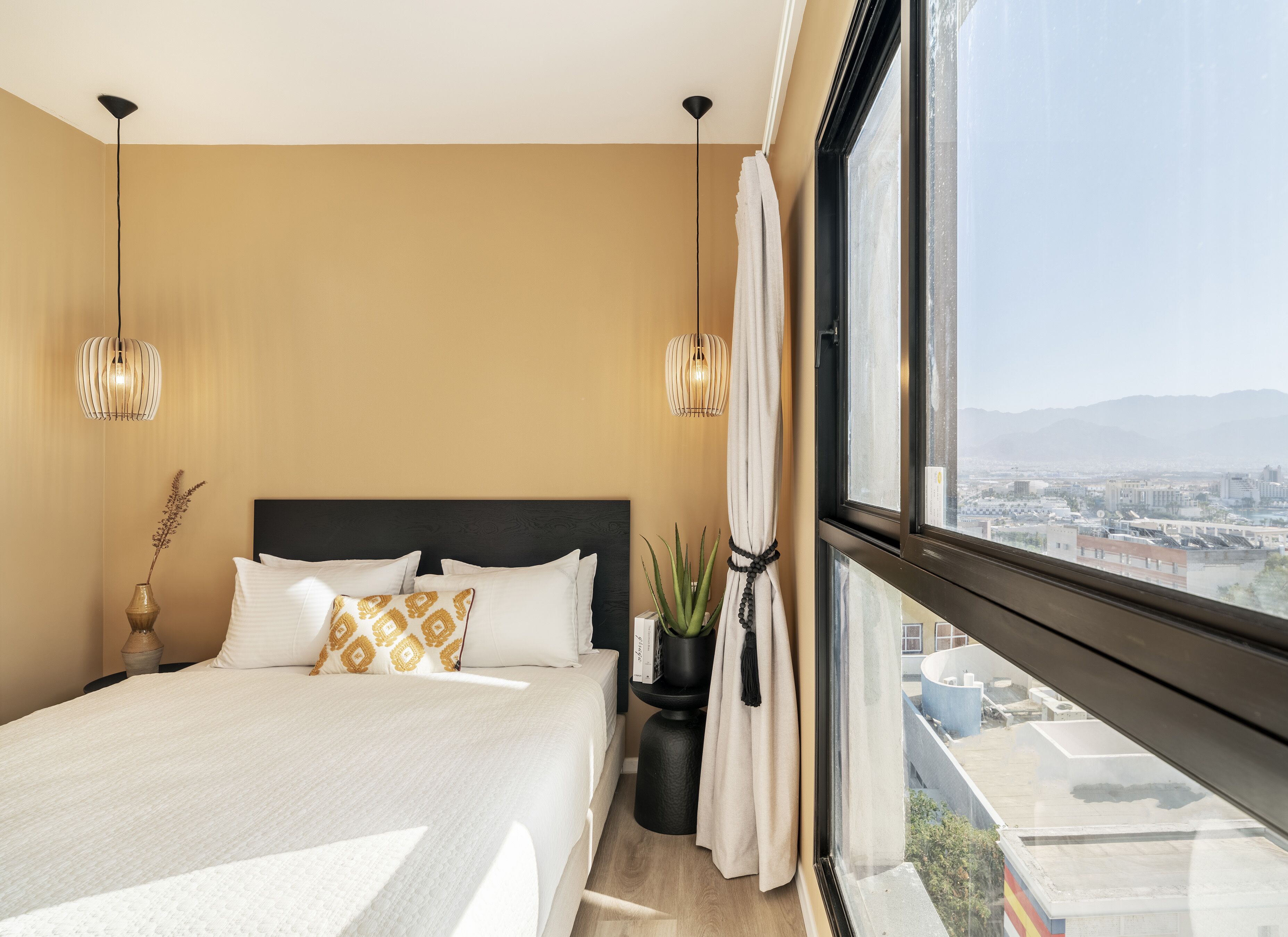 Un dormitori modern amb un llit gran, dues làmpades penjants, vista a la ciutat i una finestra amb cortines.