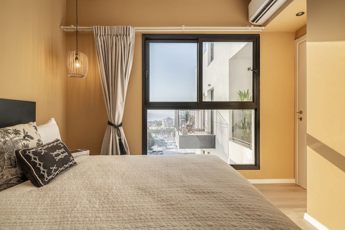 Une chambre avec un grand lit, une fenêtre donnant sur des immeubles, un luminaire suspendu et une porte.