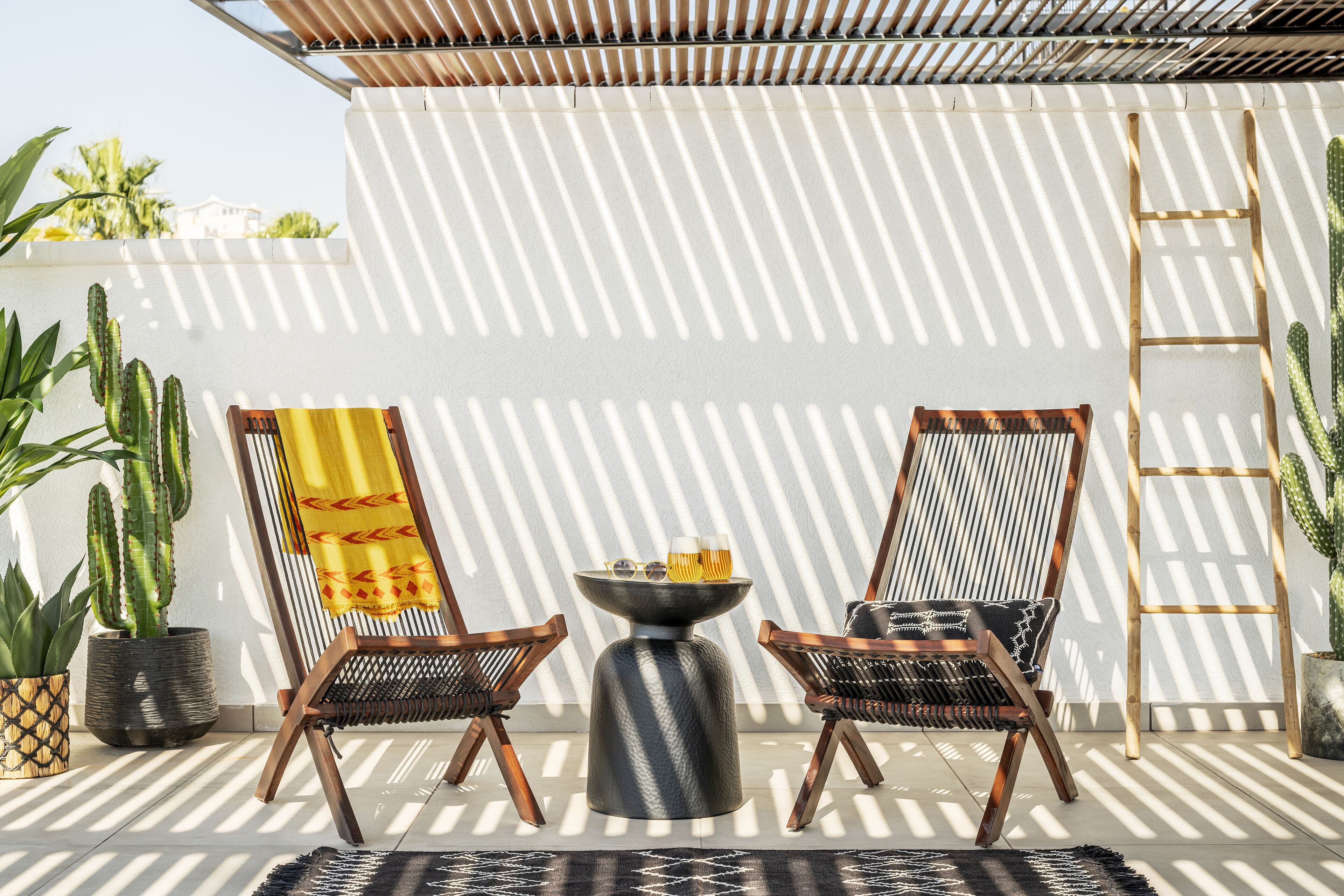 Eine Terrasse mit zwei Korbstühlen, einem kleinen runden Tisch mit Getränken und einer an einer weißen Wand stehenden Leiter, wobei Schatten sichtbar sind.