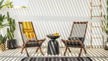 Eine Terrasse mit zwei Korbstühlen, einem kleinen runden Tisch mit Getränken und einer an einer weißen Wand stehenden Leiter, wobei Schatten sichtbar sind.